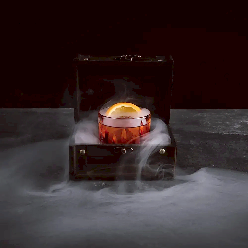 A dark alcoholic drink with a citrus slice sitting in a box that is smoking