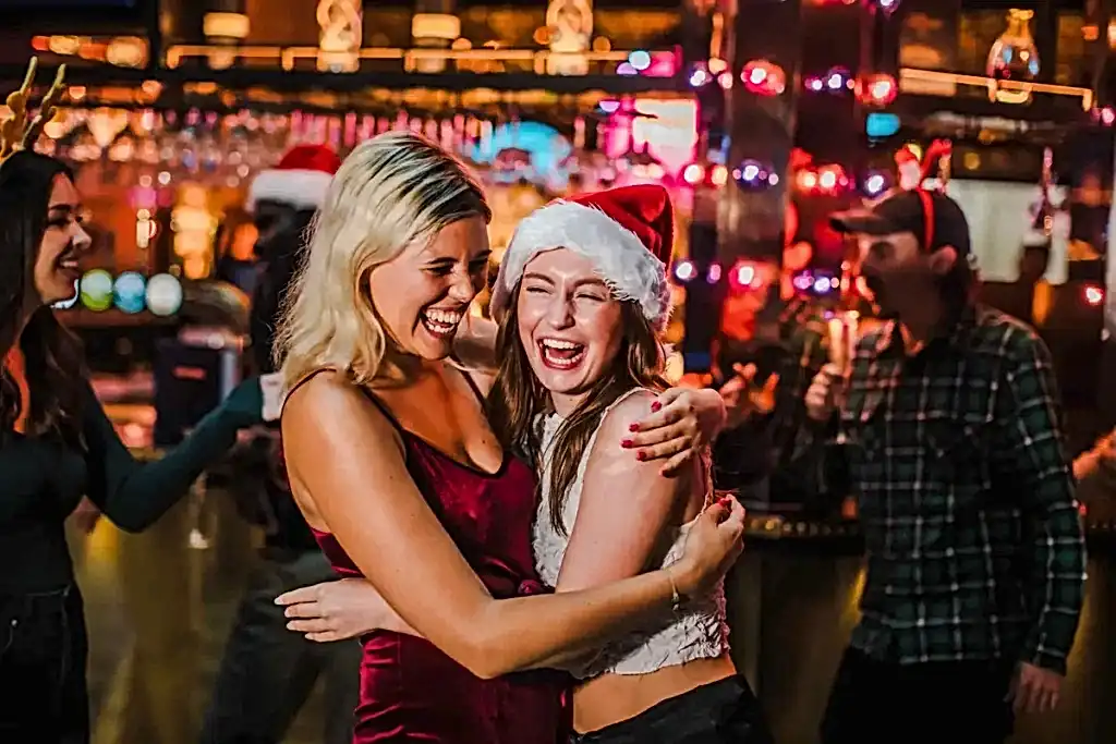 Two women wearing festive clothing, including Santa hats, hug and laugh in a lively bar setting with holiday lights in the background.