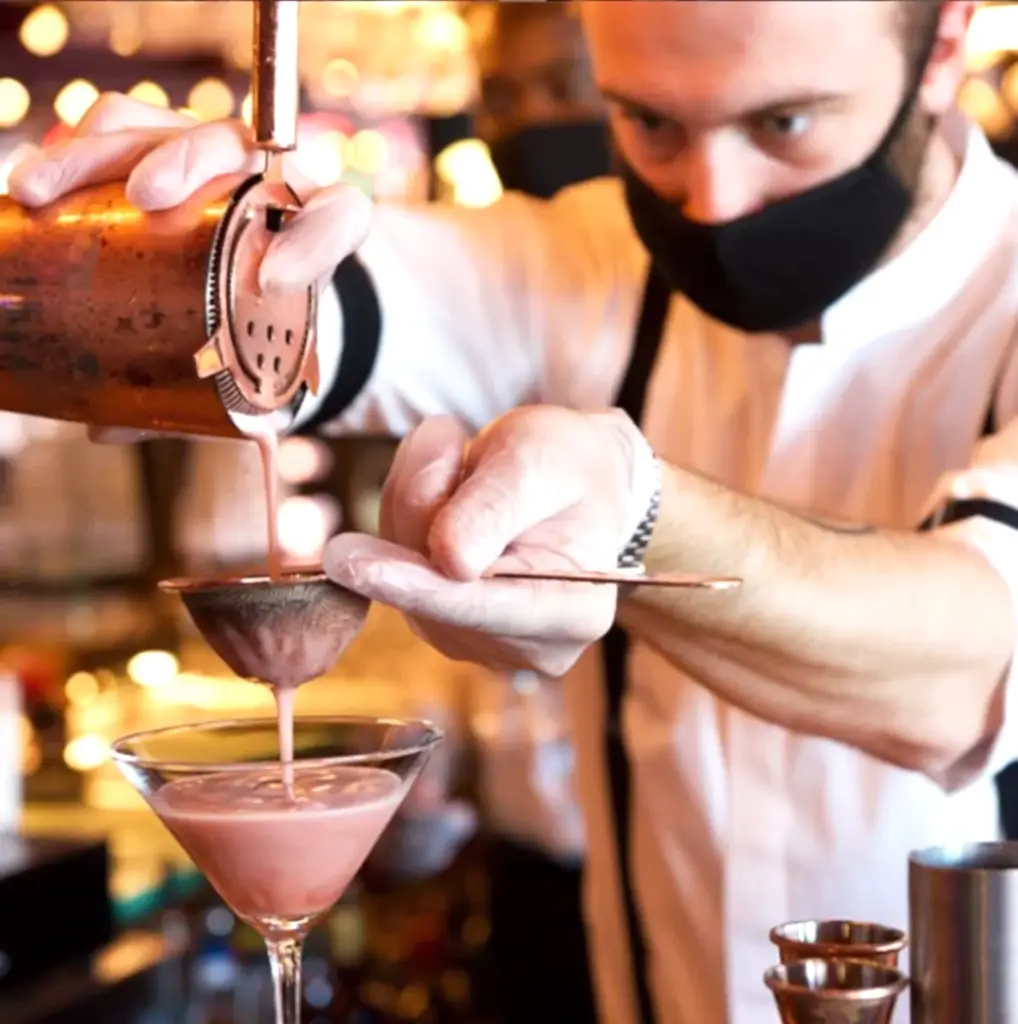 A bartender that's wearing a mask straining a cocktail into a glass
